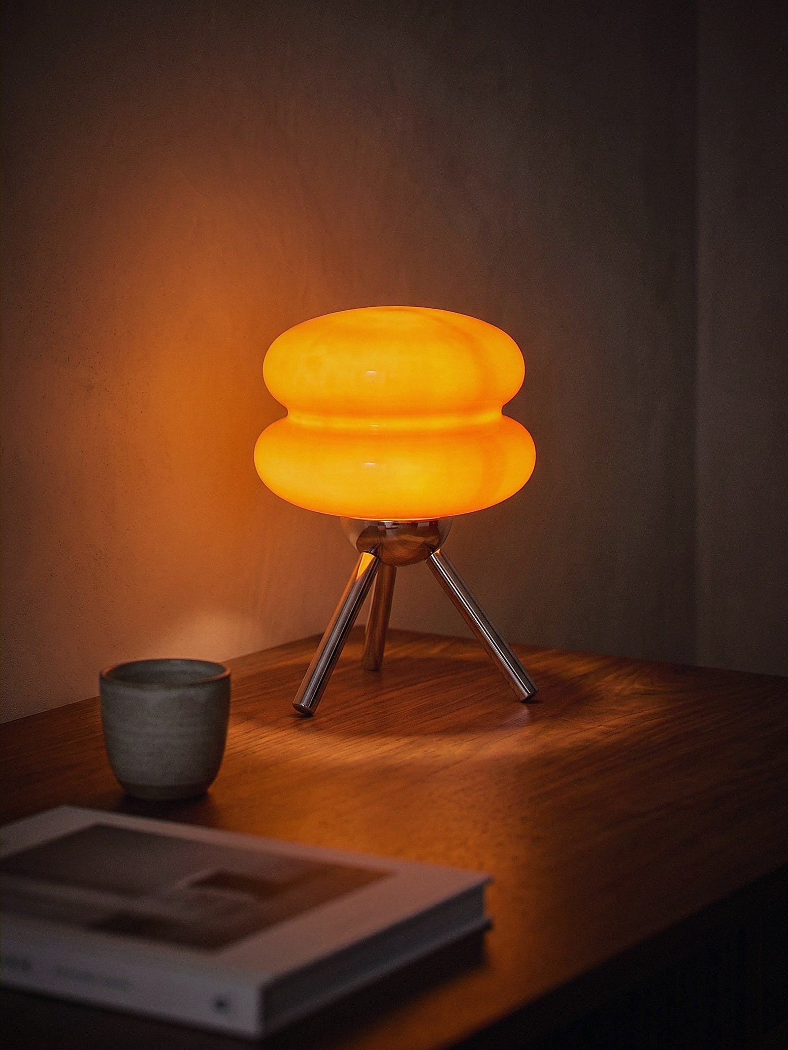 Carrara Mini, stacked orange glass rings on a polished chrome tripod, switched on and glowing warm on a small dark desk at night