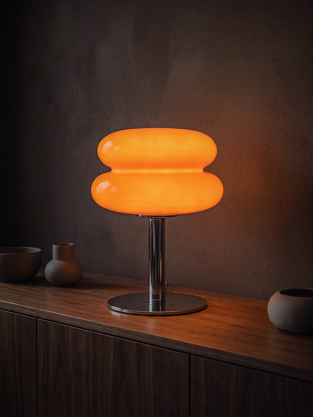 Carrara, two stacked orange glass rings on a polished chrome stem with a round chrome base, switched on and glowing warm on a dark walnut dining sideboard at night