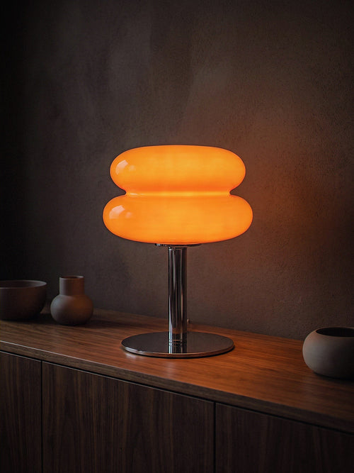 Carrara, two stacked orange glass rings on a polished chrome stem with a round chrome base, switched on and glowing warm on a dark walnut dining sideboard at night