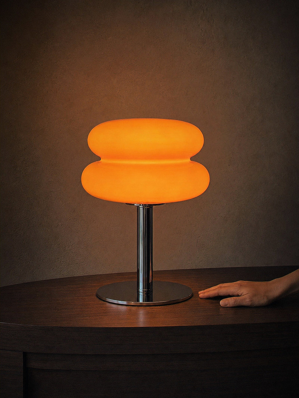 A hand resting beside Carrara, two stacked orange glass rings on a polished chrome stem with a round chrome base, showing its true size