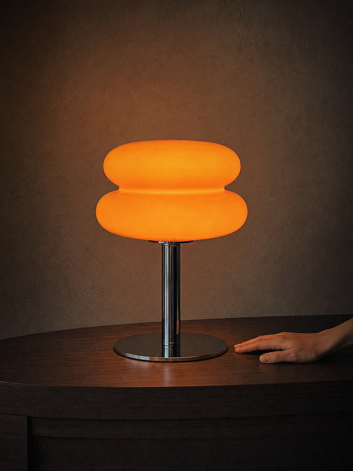 A hand resting beside Carrara, two stacked orange glass rings on a polished chrome stem with a round chrome base, showing its true size