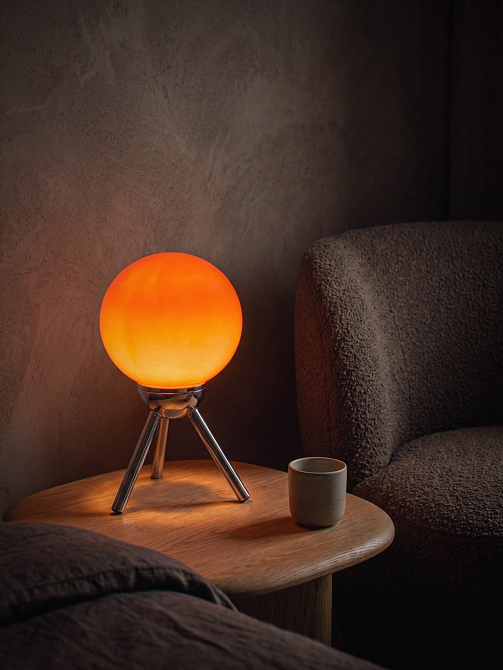 Fjord, a red-orange glass sphere held in a chrome cradle on a three-legged chrome tripod, switched on and glowing warm on a snug oak side table at night