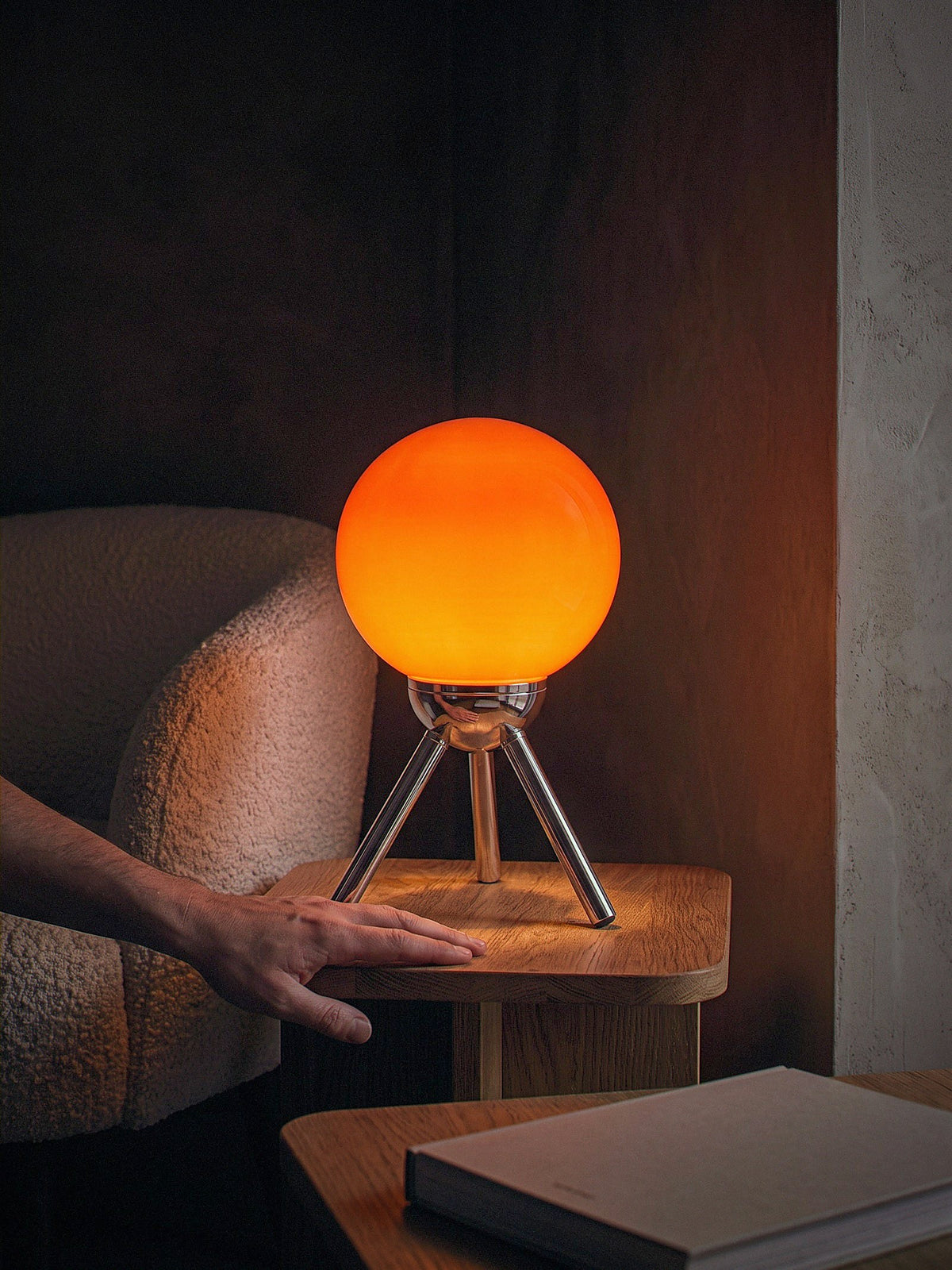 A hand resting beside Fjord, a red-orange glass sphere held in a chrome cradle on a three-legged chrome tripod, showing its true size