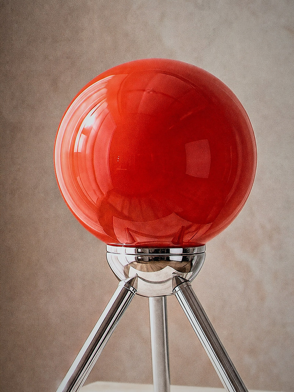Close detail of Fjord, a red-orange glass sphere held in a chrome cradle on a three-legged chrome tripod