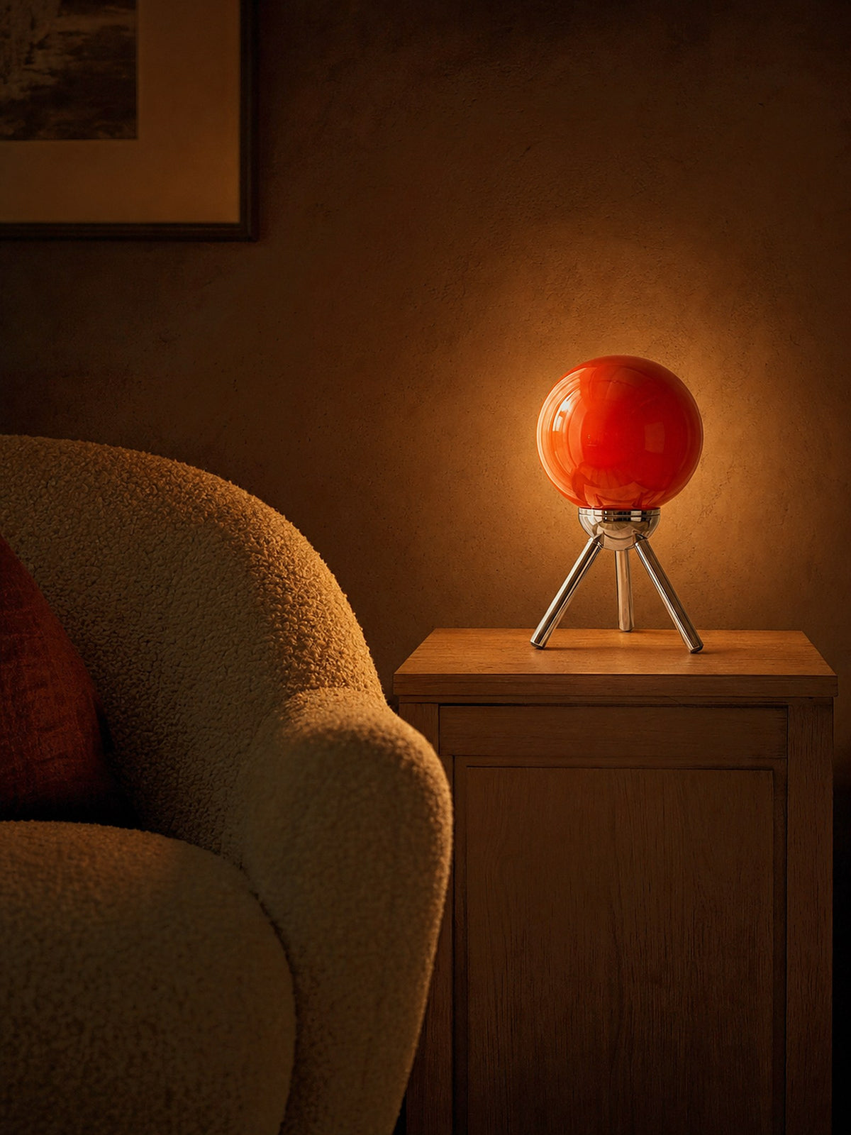 Fjord, a red-orange glass sphere held in a chrome cradle on a three-legged chrome tripod, lit in a snug reading corner