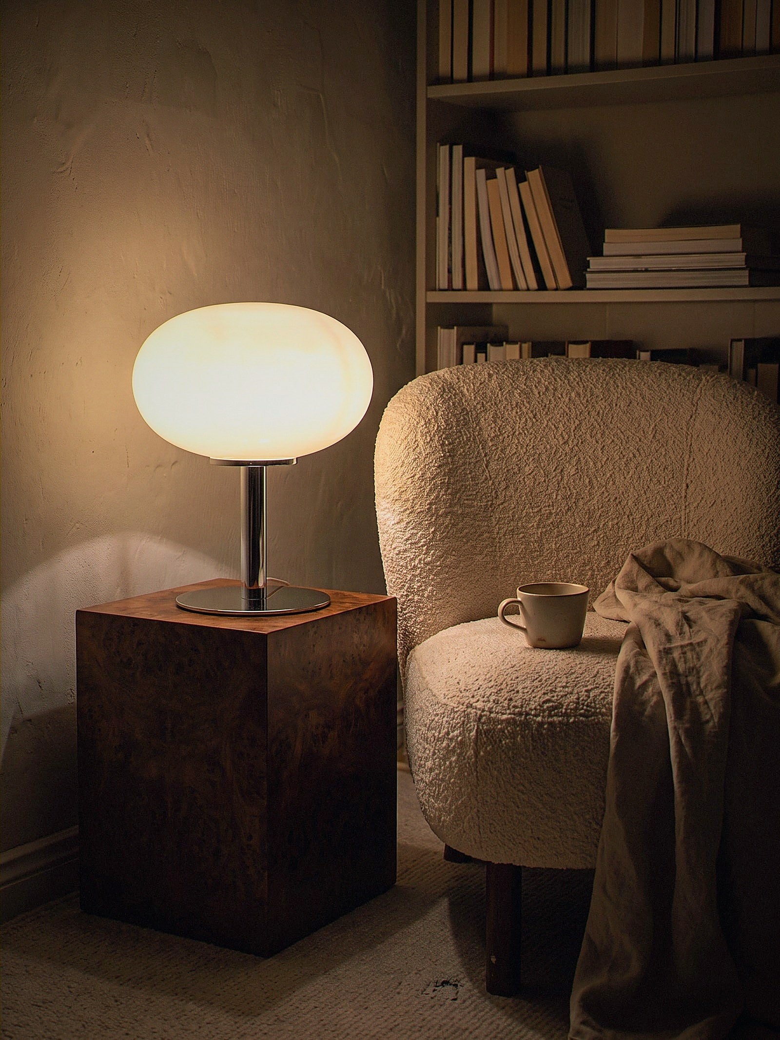 Halo, a soft cream frosted glass dome on a slim polished chrome base, switched on and glowing warm in a small library corner at night