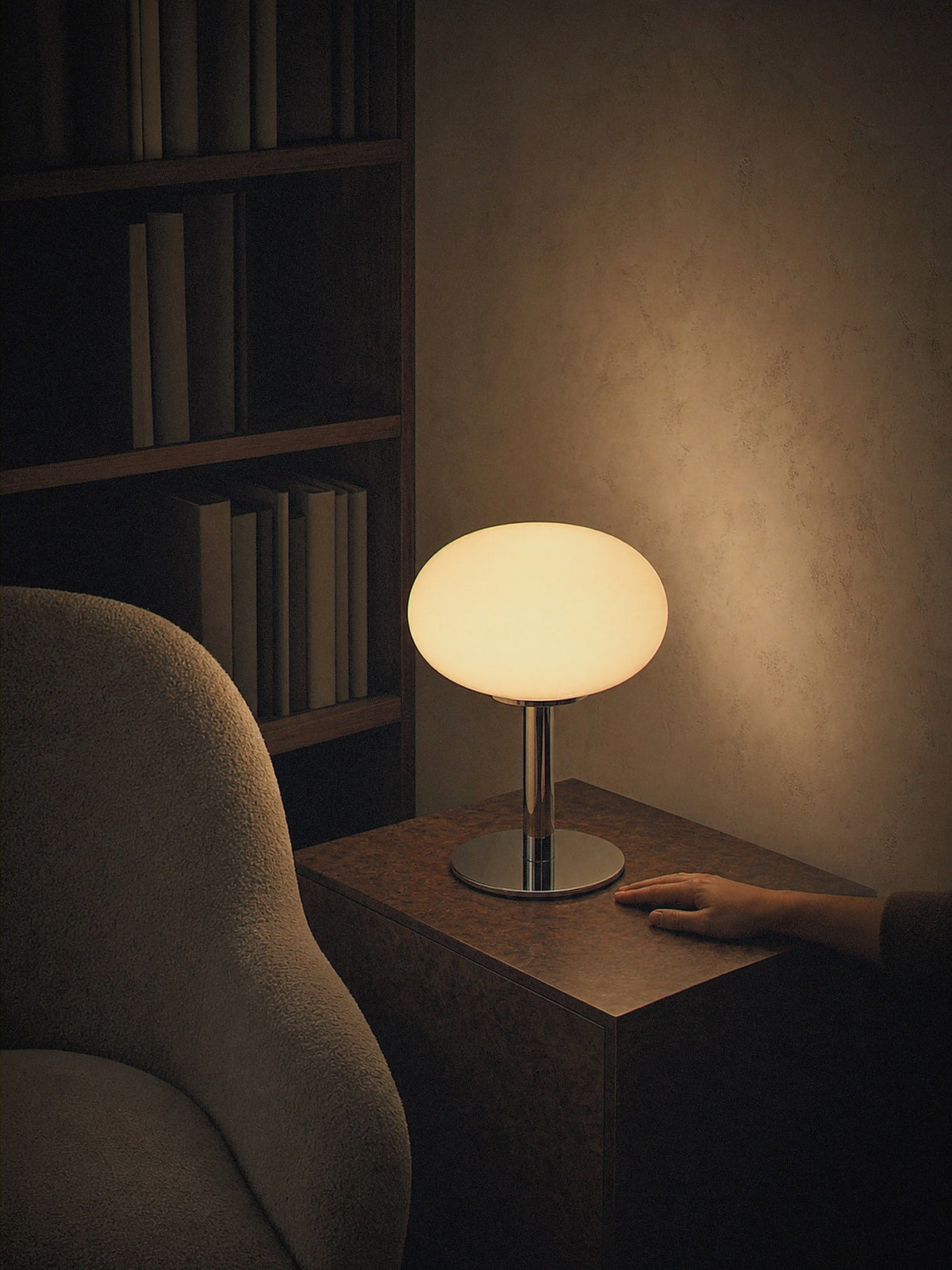 A hand resting beside Halo, a soft cream frosted glass dome on a slim polished chrome base, showing its true size