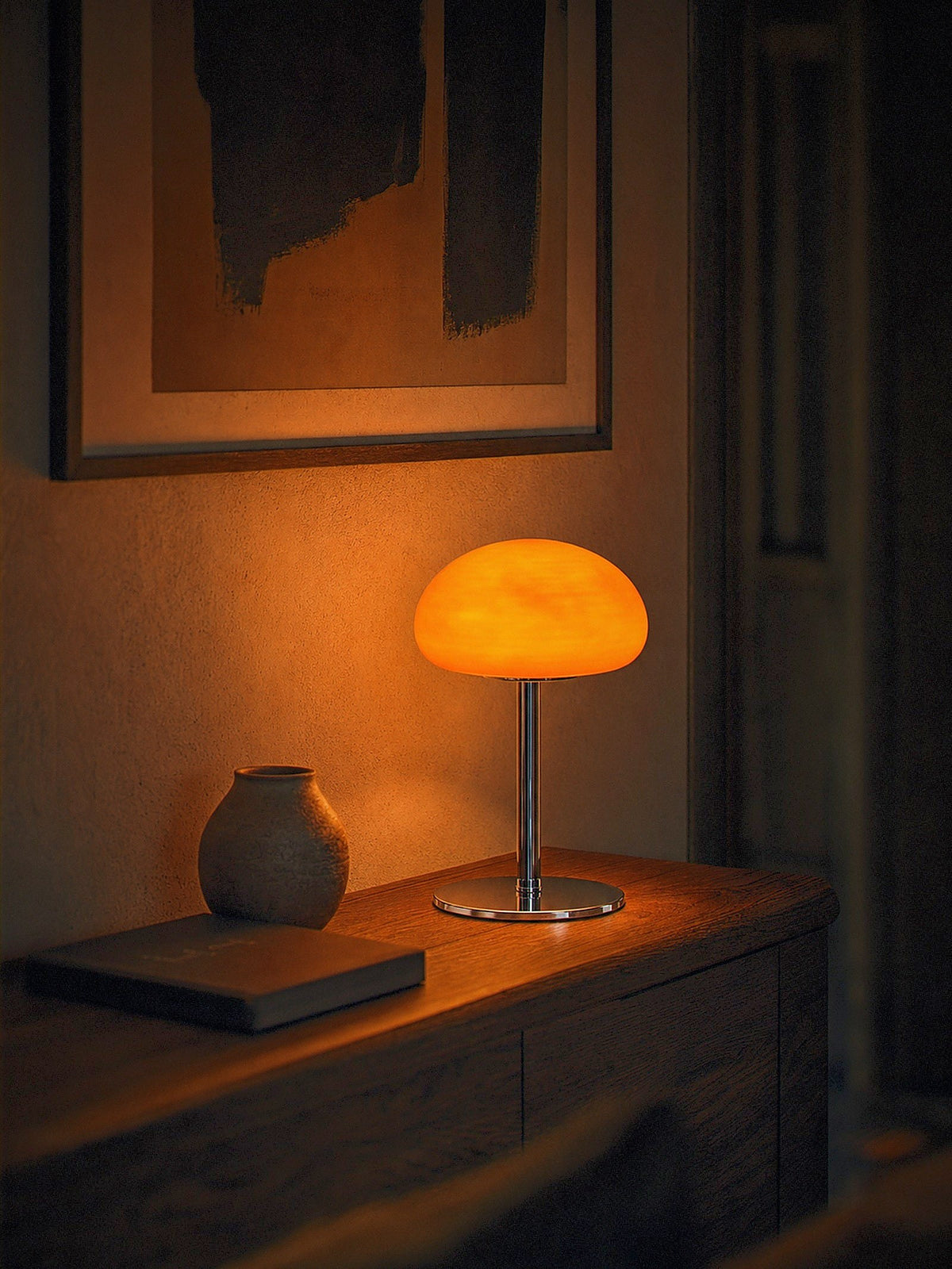 Nimbus, an orange glass dome shade on a slim chrome stem and round chrome base, switched on and glowing warm on a narrow hallway console at night