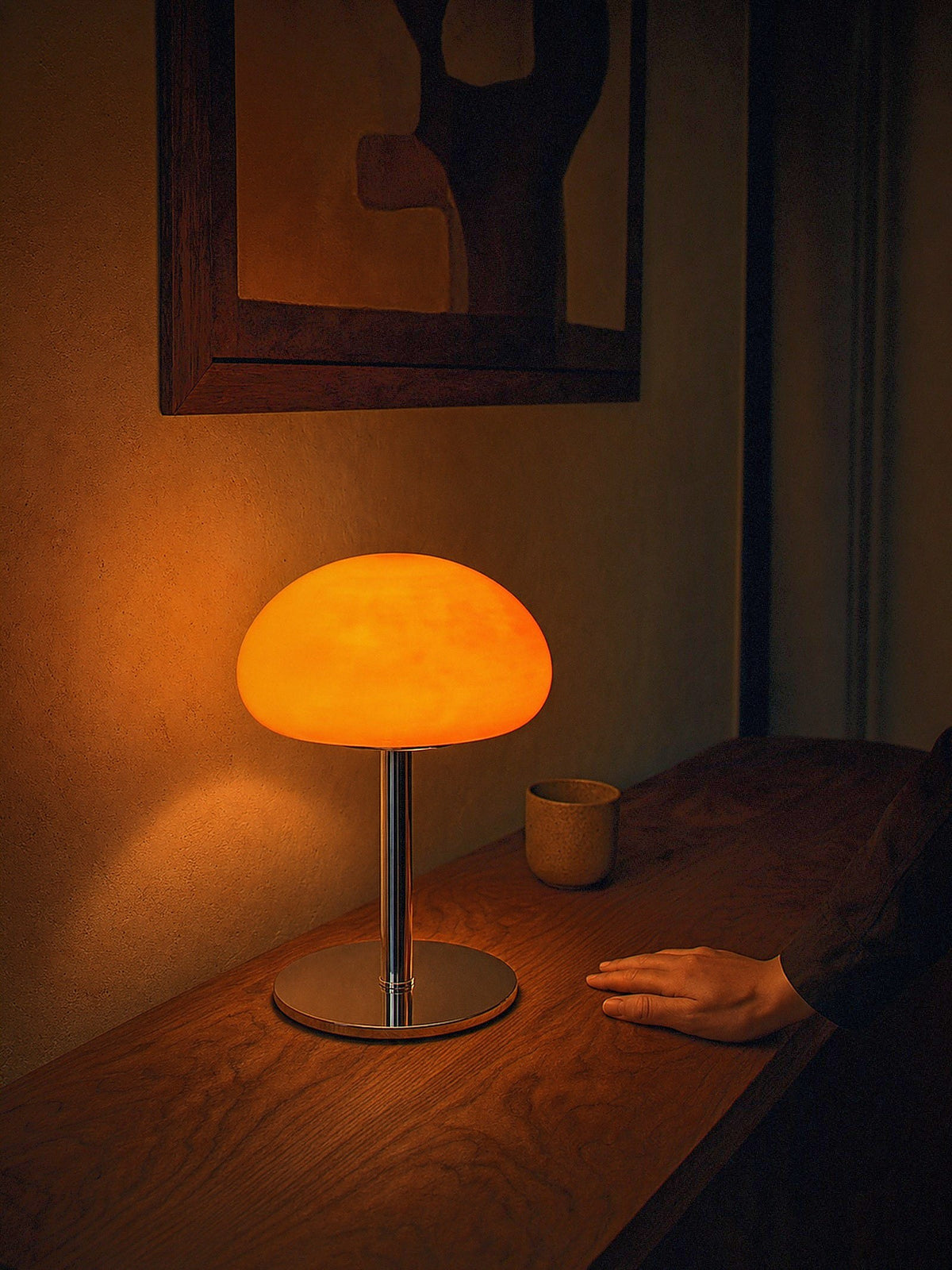 A hand resting beside Nimbus, an orange glass dome shade on a slim chrome stem and round chrome base, showing its true size