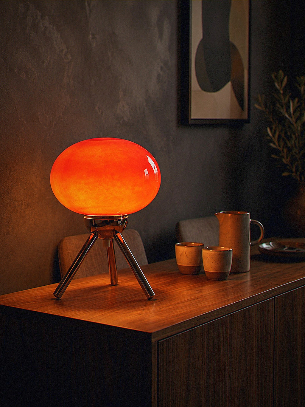 Prism, a red glass dome lamp on a polished chrome tripod, switched on and glowing warm on a dark walnut living-room console at night
