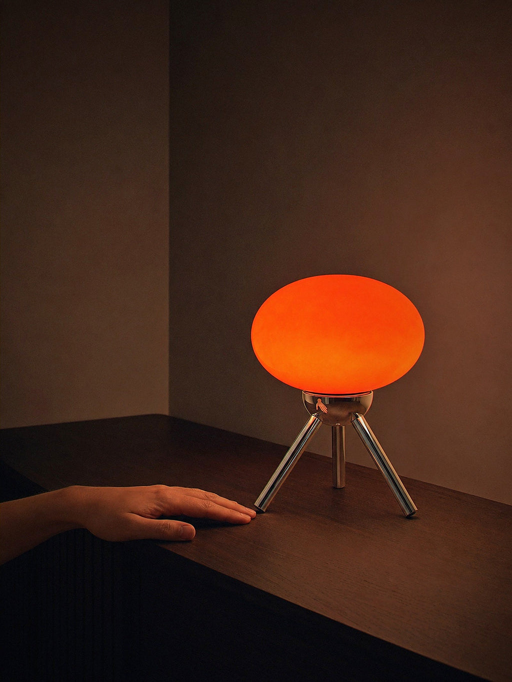 A hand resting beside Prism, a red glass dome lamp on a polished chrome tripod, showing its true size