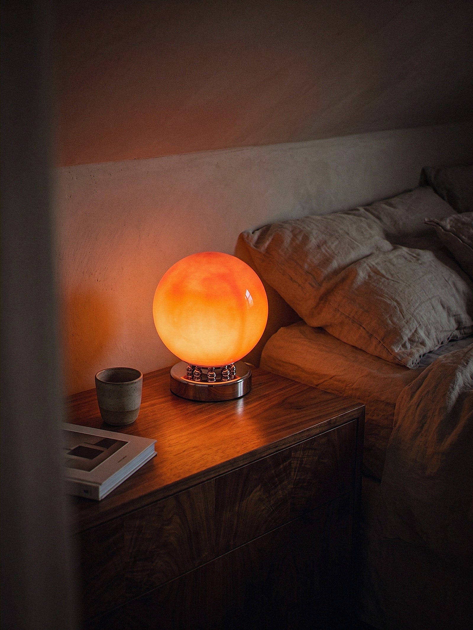 Whisper, an orange glass sphere on a chrome base ringed with polished chrome spheres, switched on and glowing warm on a dark wood bedside table at night
