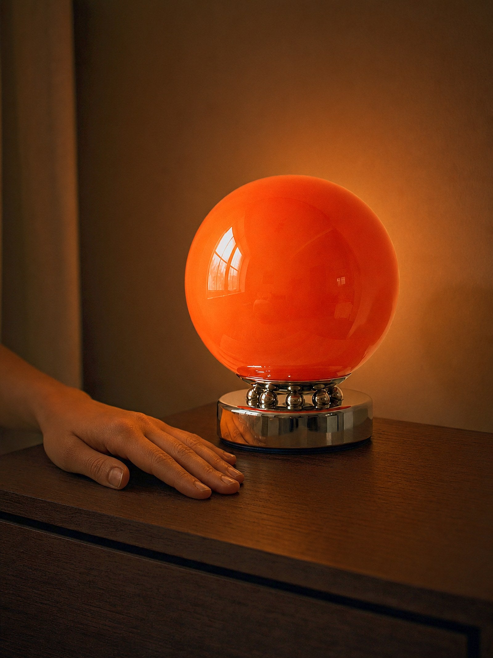 A hand resting beside Whisper, an orange glass sphere on a chrome base ringed with polished chrome spheres, showing its true size