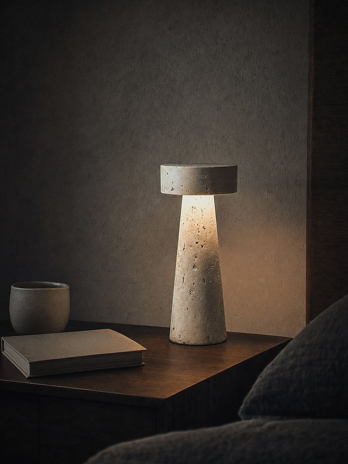 Zen, a tapered travertine column topped with a stone disc, switched on and glowing warm on a calm bedside surface at night